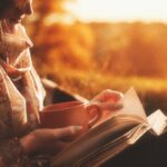 Woman in scarf reading a novel with cup in hand outside and sunset in background.